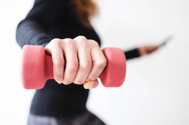 Evde fitness ve internet koçluğu konsepti, spor kıyafetleri içindeki kadının elinde dambıl ve akıllı telefon tutarak spor videosu ya da internetteki rutini takip etmesi.