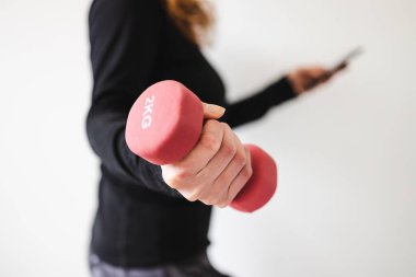 Evde fitness ve internet koçluğu konsepti, spor kıyafetleri içindeki kadının elinde dambıl ve akıllı telefon tutarak spor videosu ya da internetteki rutini takip etmesi.