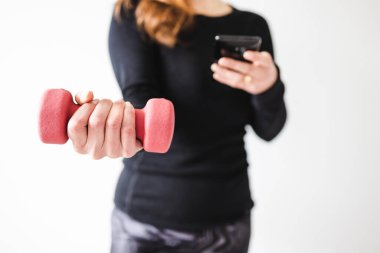 Evde fitness ve internet koçluğu konsepti, spor kıyafetleri içindeki kadının elinde dambıl ve akıllı telefon tutarak spor videosu ya da internetteki rutini takip etmesi.