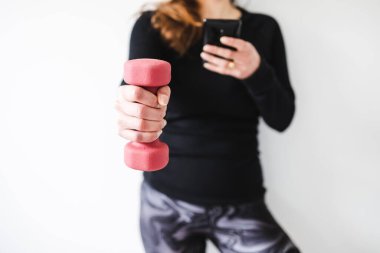 Evde fitness ve internet koçluğu konsepti, spor kıyafetleri içindeki kadının elinde dambıl ve akıllı telefon tutarak spor videosu ya da internetteki rutini takip etmesi.