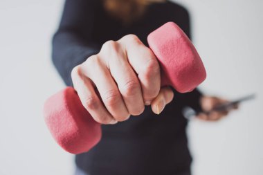 Evde fitness ve internet koçluğu konsepti, spor kıyafetleri içindeki kadının elinde dambıl ve akıllı telefon tutarak spor videosu ya da internetteki rutini takip etmesi.