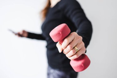 Evde fitness ve internet koçluğu konsepti, spor kıyafetleri içindeki kadının elinde dambıl ve akıllı telefon tutarak spor videosu ya da internetteki rutini takip etmesi.