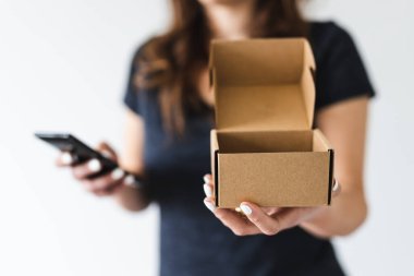 Ev teslimatı ve online alışveriş kavramı. Kadının kameraya doğru elinde teslimat paketi var. Akıllı telefonuyla sığ bir alanda fotoğraf çekiyor.