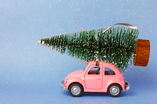 Pink toy retro car  delivering a Christmas tree  on a beautiful blue snow background.  Christmas card concept