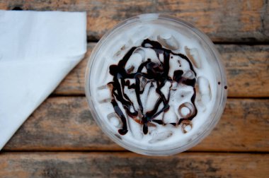 Üstünde çikolata sosu ve kağıt mendil olan taze buzlu süt dolu plastik bardağın en üst görüntüsü..