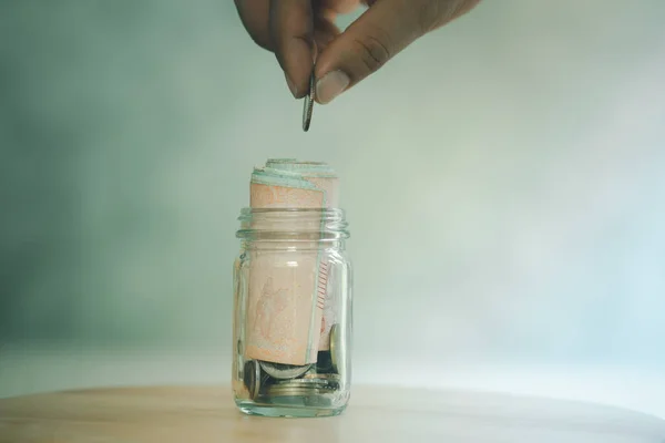 Man hand drops Thai money into a glass jar for a savings