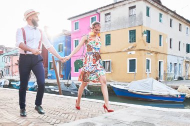 Ada sokaklarında genç ve mutlu çift Burano yürüme. Renkli elbiseli kadın, kırmızı ayakkabılar ve kafa bandı ve yeşil hasır çanta ile beyaz gömlek, hasır şapka ve koyu pantolon askısı ile adam 