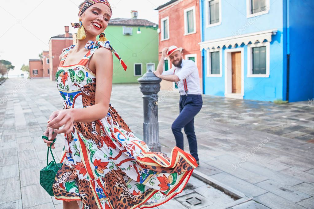 Young couple on colorful Burano island streets. In front woman in colorful dress, red shoes and with headband, man in white shirt, straw hat and dark pants with suspenders 