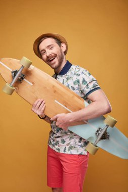Gidon bıyıklı karizmatik genç adam longboard üzerinde başka bir el longboard eliyle işaret tropikal yazdırma polo gömlek, kırmızı şort ve hasır şapka giymiş. Stüdyo sarı arka plan ile vurdu 