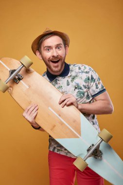 Gidon Bıyıklı heyecanlı genç adam onun elinde longboard ile tropikal yazdırma polo gömlek, kırmızı şort ve hasır şapka giymiş. Stüdyo sarı arka plan ile vurdu 