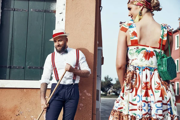 Beautiful smiling woman in colorful dress, headband and red heels and man with a broom, dressed in polka dot shirt, linen hat, bow tie and dark pants with suspenders, near colorful houses in Burano Island, Venice, Italy