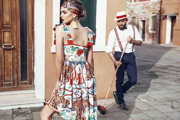 Beautiful sexy woman in colorful dress, headband and red heels and man with a broom, dressed in polka dot shirt, linen hat, bow tie and dark pants with suspenders looks at woman, near colorful houses in Burano Island, Venice, Italy