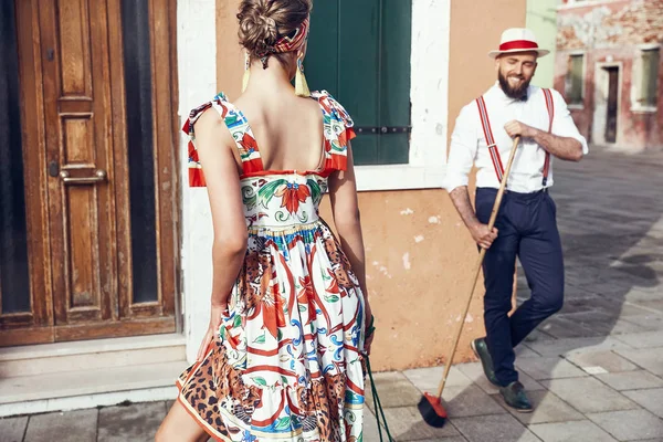 Woman in colorful dress, headband and red heels and man with a broom, dressed in polka dot shirt, linen hat, bow tie and dark pants with suspenders looks at woman, near colorful houses in Burano Island, Venice, Italy