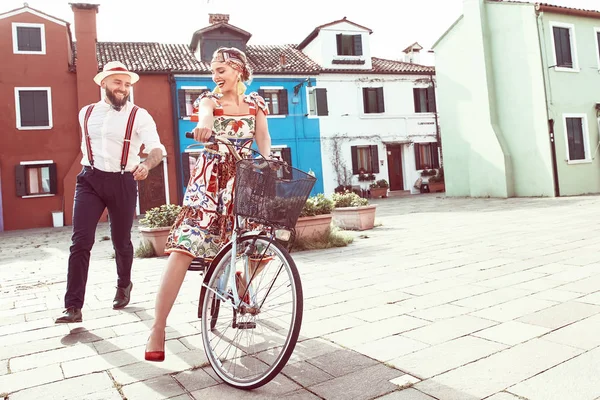 在意大利威尼斯布拉诺岛的五颜六色的房子附近 幸福的笑脸情侣 妇女礼服在多彩的浪漫礼服 头带和大耳环 骑自行车 人在白色衬衣 亚麻帽子和长裤与吊带 图库图片