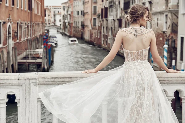 Arka, dövmeli güzel gelin giyinmiş şık, lüks Fildişi elbise ve kabarık saç modeli içinde Venedik kanal üzerinde bir köprü üzerinde poz