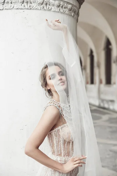 Beautiful bride in long elegant, luxury ivory dress and messy updo hairstyle, covered face by veil, posing near columns in Venice, Italy