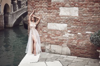 Güzel gelin çıplak ile tuğla duvara yakın poz giyinmiş şık, lüks Fildişi elbise ve dağınık kabarık saç modeli, oluşturan ve Venedik canal