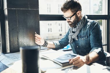 Adam mimar koyu saç ve sakal, kot ceket, eşarp ve gözlük sahte pencerenin oturuyor ve yeni bir proje, elinde kalem ile tasarımlar