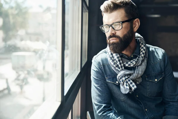 Pensive young man with dark hair and beard, weared in denim jacket, scarf and eyeglasses is sitting near big window