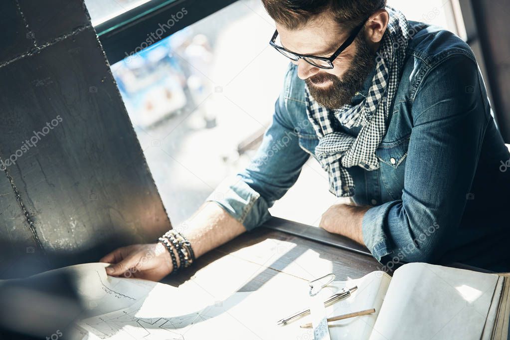 Man architect with dark hair and beard, weared in denim jacket, scarf, eyeglasses is sitting in the modern office near the window and designs a new project