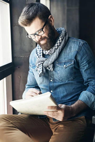 Smiling male writer journalist with dark hair and beard in denim jacket and eyeglasses is sitting in public place and creating new article