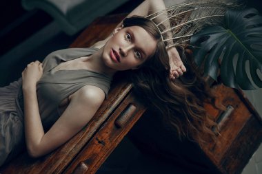 Taze makyaj, kuş tüyü taç olan kadın büyülü yumuşak portresi. Kadın masada, büyük bitki yanında yatıyordu