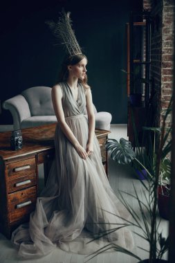 Büyülü genç kadın kafasına, tüy tacıyla uzun hafif zeytin tül elbise vintage tablo ve kamera yakın durumda