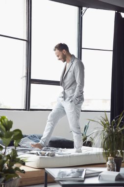 Yakışıklı adam koyu saç ve sakal hafif bir ceket ve keten pantolon İskandinav studio büyük pencere ve bitkiler ile yatakta duruyor