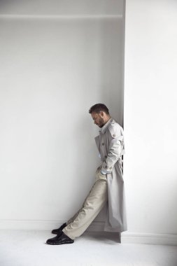 Moda portre yakışıklı adam karanlık sakal ve saç, ışık palto, gömlek, bej pantolon ve siyah ayakkabılar renksizdir. Adam duvara yakın duruyor