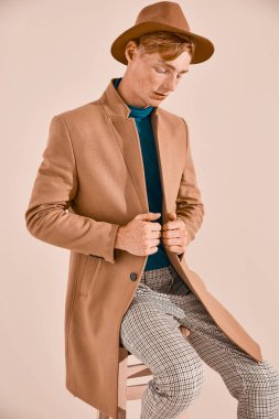 Kızıl saçlı ve çilli kahverengi palto ve şapka, ekose pantolon, Turkuaz kazak, sandalyede oturan poz ile genç erkek modeli. Pembe bir arka plan 