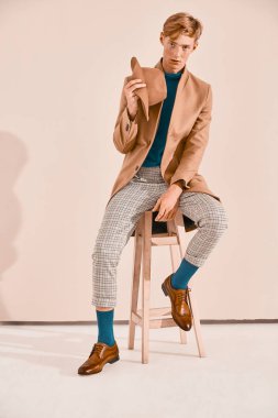Genç erkek turkuaz kazak ve sandalyede oturan çorap, kahverengi ayakkabı, Kızıl saçlı ve çilli ekose pantolon, kahverengi ceket içinde poz ile model. Pembe bir arka plan 