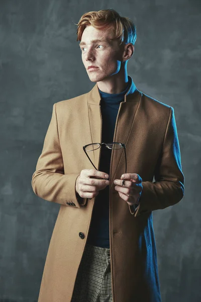 Studio portrait of young male model with red hair and freckles. Weared in brown coat, plaid pants, with glasses in hand. Blue light, gray background.