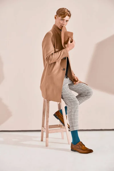 Young male model with red hair and freckles in brown coat and hat, plaid pants, turquoise sweater and socks, brown shoes, sitting on chair. Pink background