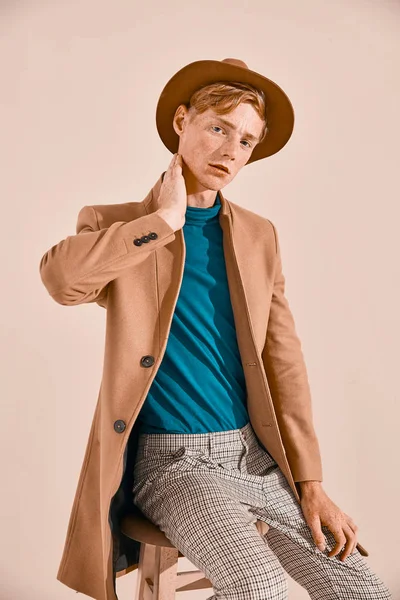 赤い髪とそばかすのポーズ茶色のコートと帽子格子縞のズボンを椅子に座って ターコイズ ブルーのセーターの若い男性モデル ピンクの背景 — ストック写真