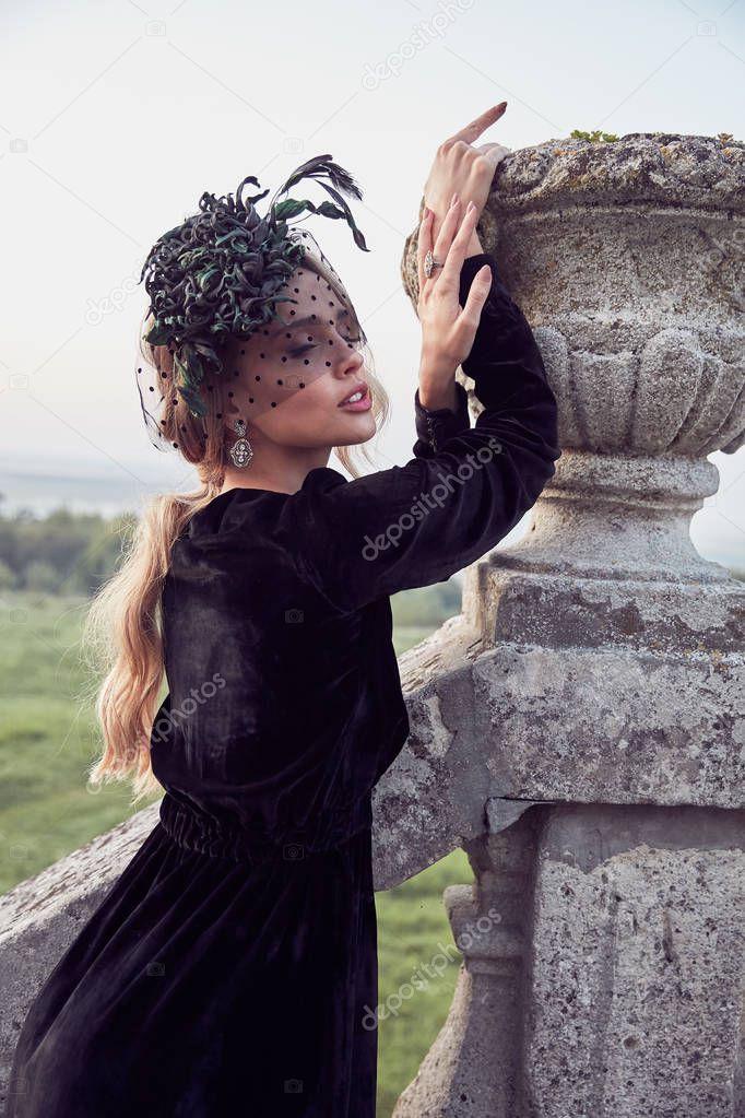 Outdoor portrait of beautiful woman near ancient flower bed. Black dress and dark green fascinator with veil. Victorian look.