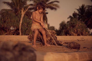 Mowgli çocuk sahilde eski ağaçta oturuyor ve kum bir sopa ile çizim, kıvırcık saçlı. Arka plan üzerinde tropikal ağaçlar