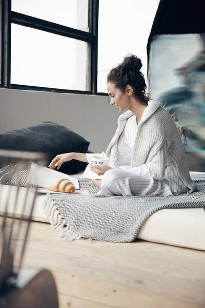 Cozy home portrait of young girl having breakfast on bed and reading magazine. Scandinavian room.