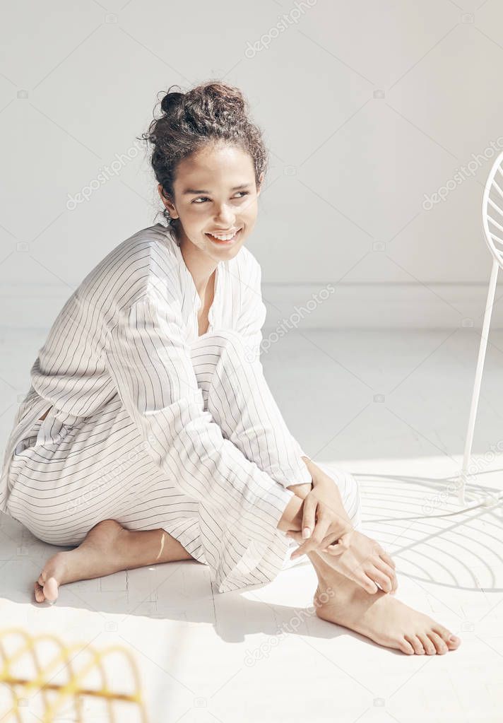 Light cozy portrait of young smiling girl wearing white pajama sitting on the floor. White room, natural lighting