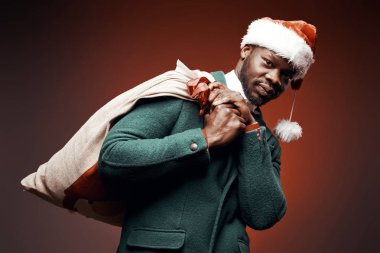 Modern Noel Baba. Yeşil ceket ve santa çantası ile kırmızı kazaklı adam elinde gülümseyerek. Ateş, kırmızı arka plan stüdyo