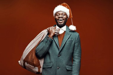 Modern Noel Baba. Yeşil ceket ve santa çantası ile kırmızı kazaklı adam elinde gülümseyerek. Ateş, kırmızı arka plan stüdyo