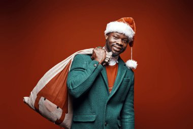 Modern Noel Baba. Yeşil ceket ve santa şapka ve çanta ile kırmızı kazaklı poz gülümseyen duygusal adam. Ateş, kırmızı arka plan stüdyo