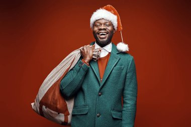Modern Noel Baba. Yeşil ceket ve santa şapka ve çanta ile kırmızı kazaklı poz gülümseyen duygusal adam. Ateş, kırmızı arka plan stüdyo