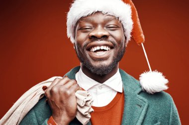 Modern Noel Baba. Yeşil ceket ve santa şapka ve çanta ile kırmızı kazaklı poz gülümseyen duygusal adam. Ateş, kırmızı arka plan stüdyo