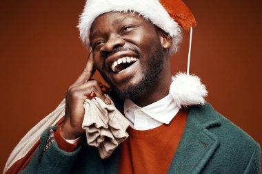 Modern Noel Baba. Yeşil ceket ve santa şapka ve çanta ile kırmızı kazaklı poz gülümseyen duygusal adam. Atış, kahverengi zemin stüdyo