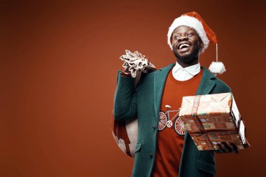 Modern Noel Baba. Yeşil ceket ve santa şapka ve çanta ve hediye ile kırmızı kazaklı poz gülümseyen duygusal adam. Atış, kahverengi zemin stüdyo