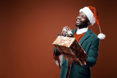 Modern Noel Baba. Yeşil ceket ve santa şapka ve çanta ve hediye ile kırmızı kazaklı poz gülümseyen duygusal adam. Atış, kahverengi zemin stüdyo