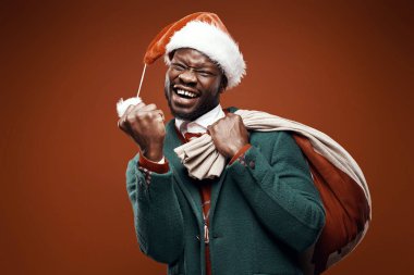 Modern Noel Baba. Yeşil ceket ve santa şapka ve çanta ile kırmızı kazaklı poz gülümseyen duygusal adam. Atış, kahverengi zemin stüdyo