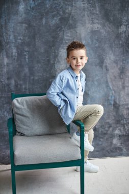 Loft studio, gri backgroung sandalyede oturan mavi gömlekli moda çocuk.