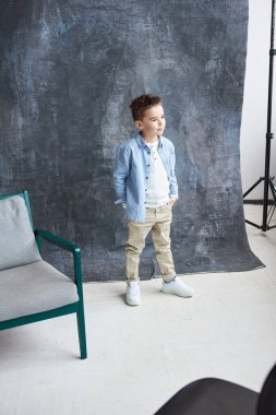 Mavi gömlek ve bej pantolon hafif çatı stüdyo büyük pencere ve gri sandalye ile yürüyüş şık çocuk