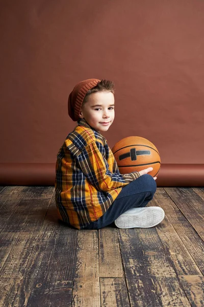 おしゃれな男の子バスケット ボール ボールの手の中で床に座って スタジオ撮影 茶色の背景 — ストック写真